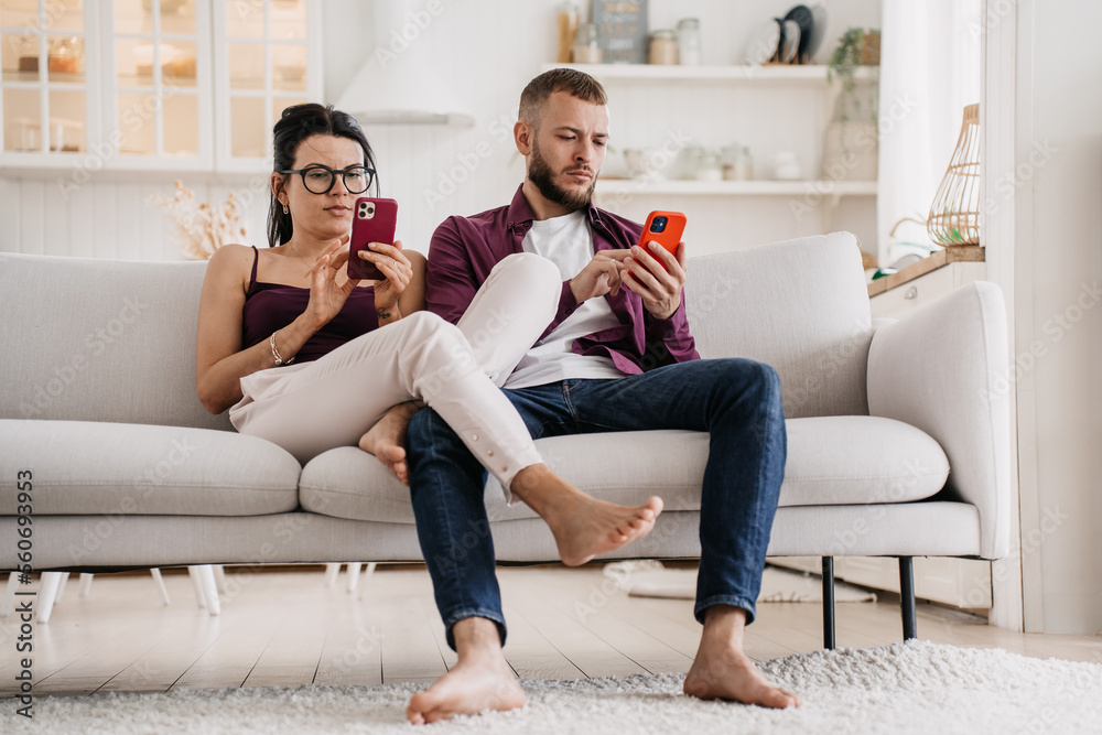 Young caucasian couple in casual sits on couch at home using phones, searching new apartment via internet. Relocation. Serious man at social media. Changing real life for virtual. Marriage troubles. - obrazy, fototapety, plakaty 