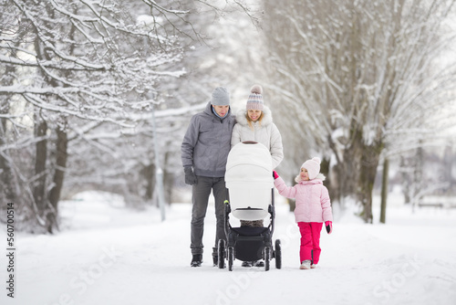 Happy young adult parents and little daughter pushing white baby stroller and walking on snow covered sidewalk at park in cold winter day. Spending time together. Two child family. Front view.