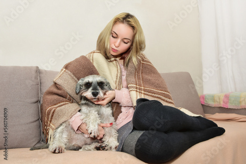A beautiful lady in a warm blanket sits comfortably on the sofa hugging her cute dog