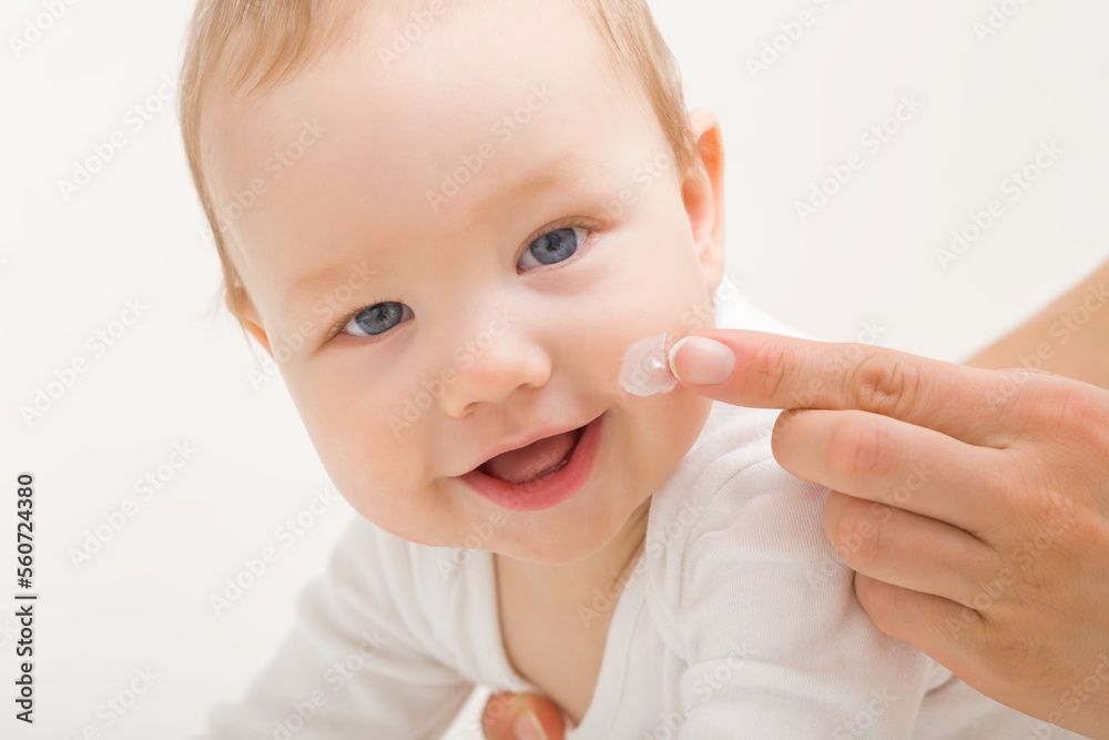 Young adult mother finger applying white moisturizing cream on baby boy cheek on light gray background. Daily care about face skin. Closeup. Front view. Happy smiling infant. Young adult mother finger applying white moisturizing cream on baby boy cheek on light gray background. Daily care about face skin. Closeup. Front view. Happy smiling infant.