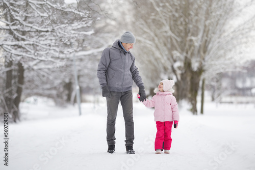 Happy smiling little daughter and young adult father walking on white snow covered sidewalk at park. Spending time together in beautiful cold winter day. Enjoying stroll. Front view.