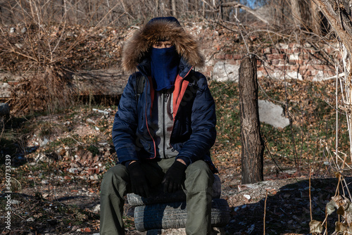 A grown man wearing a mask, wearing a hooded jacket, and wearing gloved military pants sits and looks into the camera