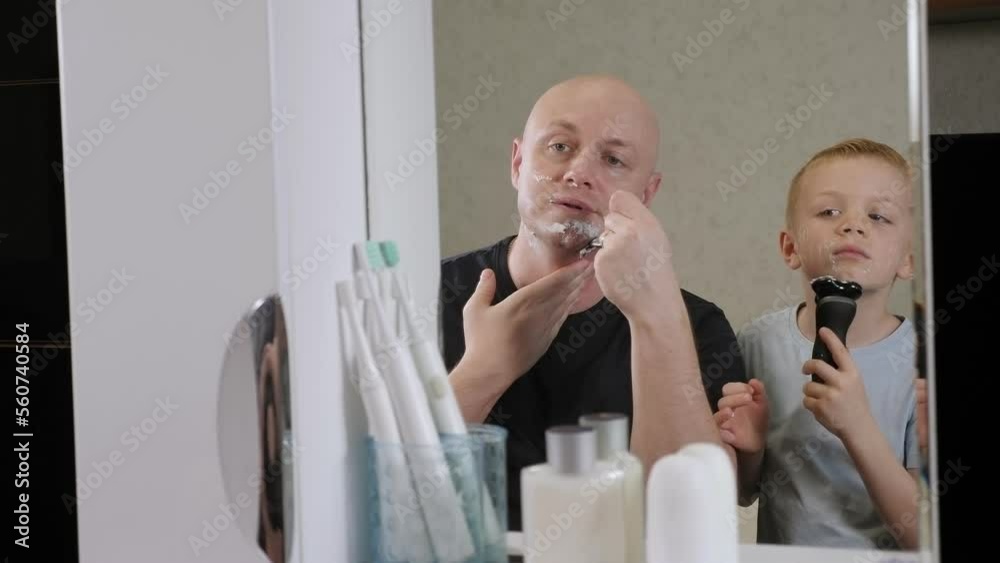 Cheerful father and son shave together in the morning in the bathroom, a boy shaves with an electric razor. Reflection in the mirror. Cheerful father and son shave together in the morning in the bathroom, a boy shaves with an electric razor. Reflection in the mirror.