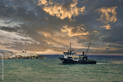 View of a fishing boat returning from the sea to land and a view of the small harbor, lighthouse and gorgeous cloudy dramatic sky.