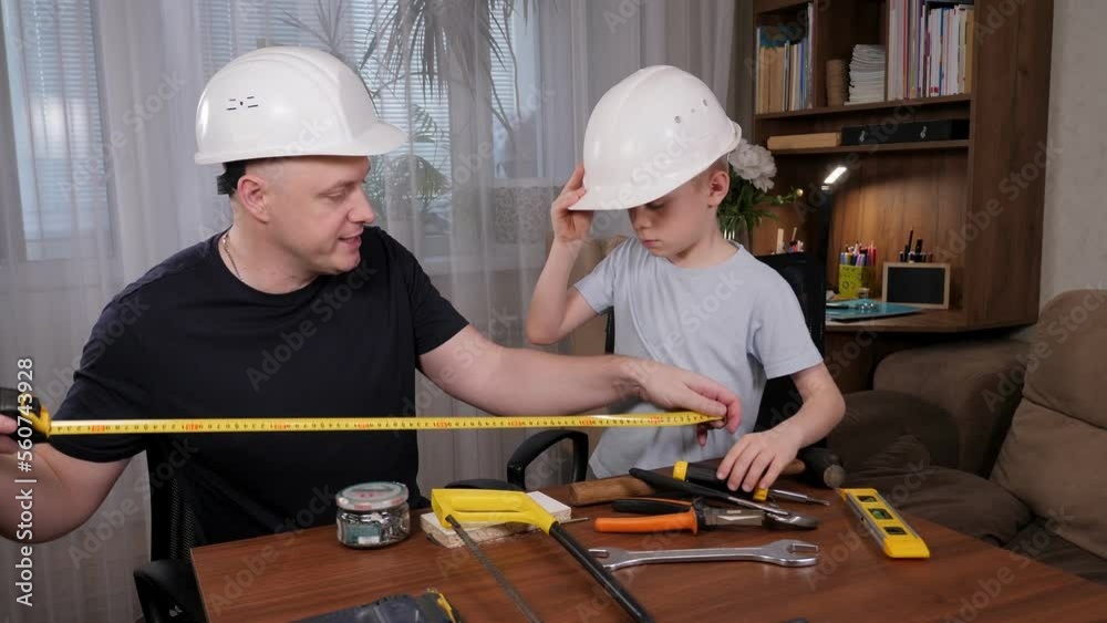 A handsome bald man shows the tools to his son while sitting at the table, the father shows how to measure the table with a tape measure.