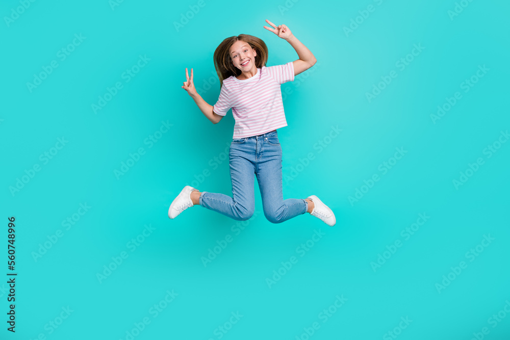 Full length photo of pretty shiny girl dressed pink t-shirt jumping high showing two v-signs isolated teal color background Full length photo of pretty shiny girl dressed pink t-shirt jumping high showing two v-signs isolated teal color background