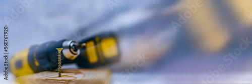 Metal self-tapping screw is screwed into the wooden board against the background of an electric screwdriver. Long banner with glow effects.