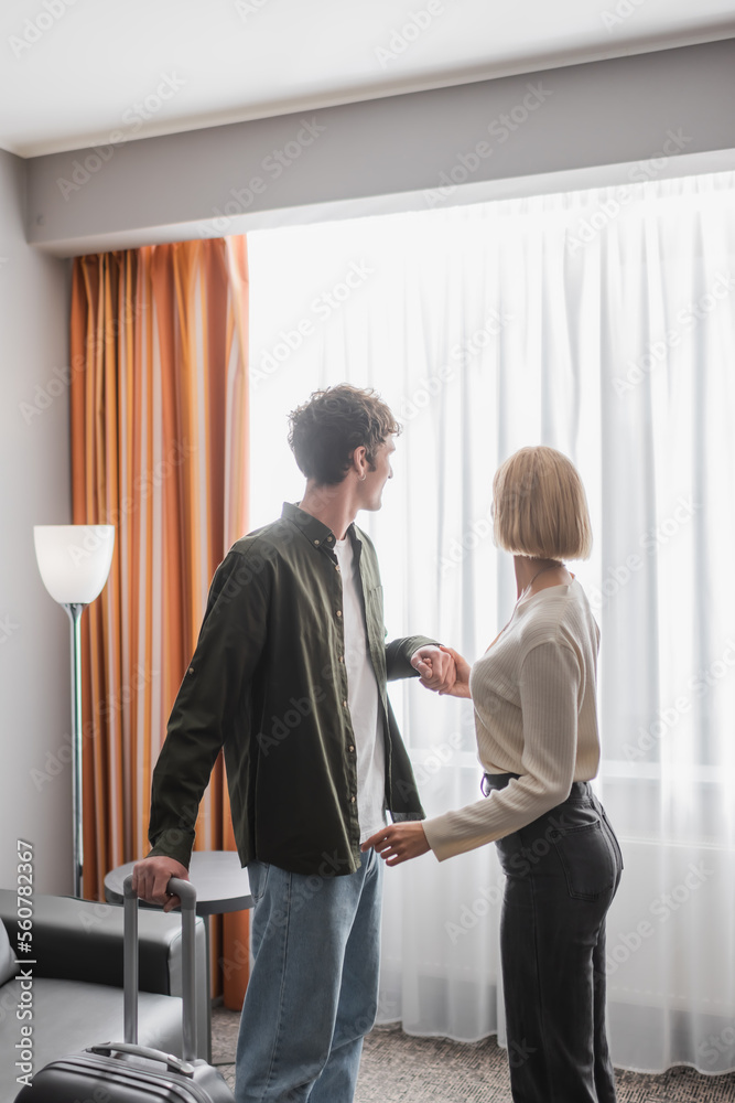young man with suitcase holding hands with girlfriend near window with curtains in hotel. young man with suitcase holding hands with girlfriend near window with curtains in hotel.