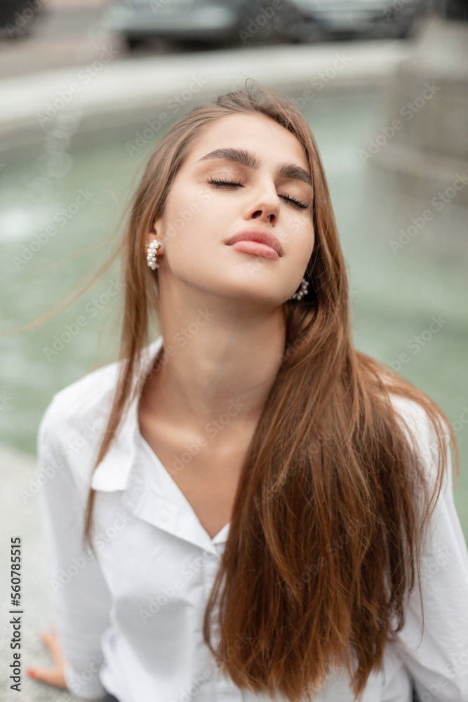 Pretty woman with nature beauty face in white shirt sits and rest on the street near a fountain Pretty woman with nature beauty face in white shirt sits and rest on the street near a fountain