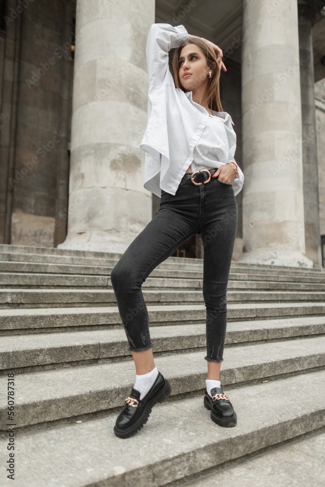 Pretty stylish young girl in fashionable business casual clothes with a white shirt, black jeans and shoes stands on the steps near a vintage building with columns Pretty stylish young girl in fashionable business casual clothes with a white shirt, black jeans and shoes stands on the steps near a vintage building with columns