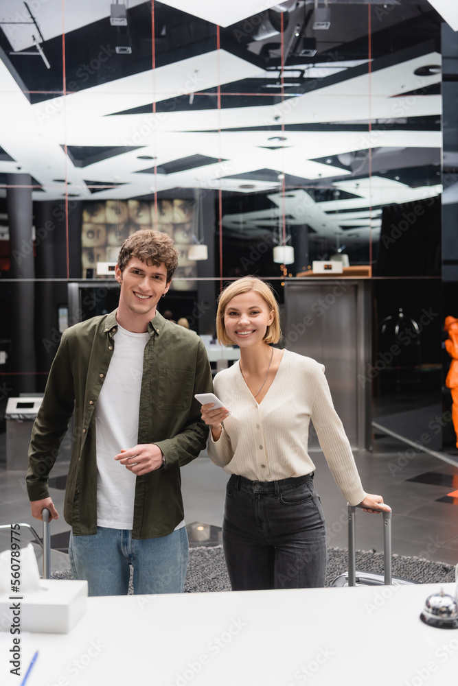 young man and cheerful blonde woman with mobile phone looking at camera near reception in modern hotel. young man and cheerful blonde woman with mobile phone looking at camera near reception in modern hotel.