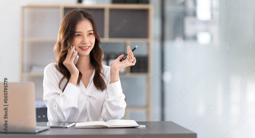 Young Asian businesswoman sit with her laptop computer happily talking on the phone with her customer explaining the detail in the office. Young Asian businesswoman sit with her laptop computer happily talking on the phone with her customer explaining the detail in the office.