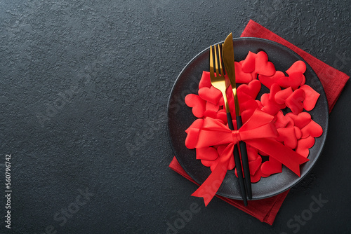 Valentines day romantic table setting. Empty or closeup of a dinner black plate, knife, fork and decorative silk hearts on black background. Holiday concept. Copy space for inscriptions.