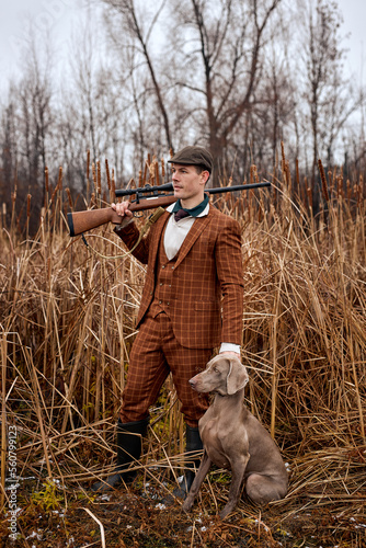 shikari with gray hunting dog in autumn forest. Image taken during big game hunting trip. Hunting period, autumn season open. handsome caucasian guy in brown suit outdoors in nature photo