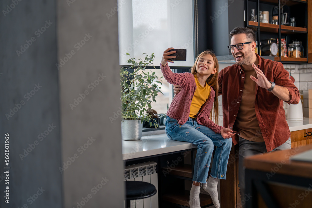 Father and daughter having a video call on a smart phone or taking a selfie. Cheerful father and his adorable girl looking at the front camera on the phone, he is waving, the both of them are smiling.