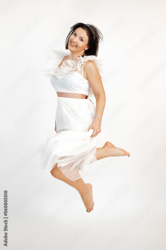 Bare footed cheerful adult brunette woman joyfully jump in airy romantic white dress, isolated on white background, copy space. Holiday look, event celebration, elegant female clothes, festive outfit