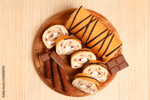 Cutting board with tasty pieces of sponge cake roll, cinnamon and chocolate on wooden background