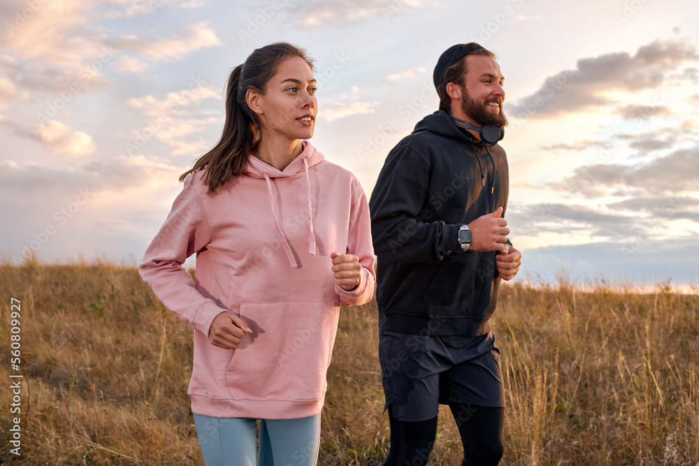 Couple in sportswear running in field. Active rest on weekends. Traveling in nature. Young caucasian male and woman in sportive outfit engaged in sport, fitness, lead healthy lifestyle.