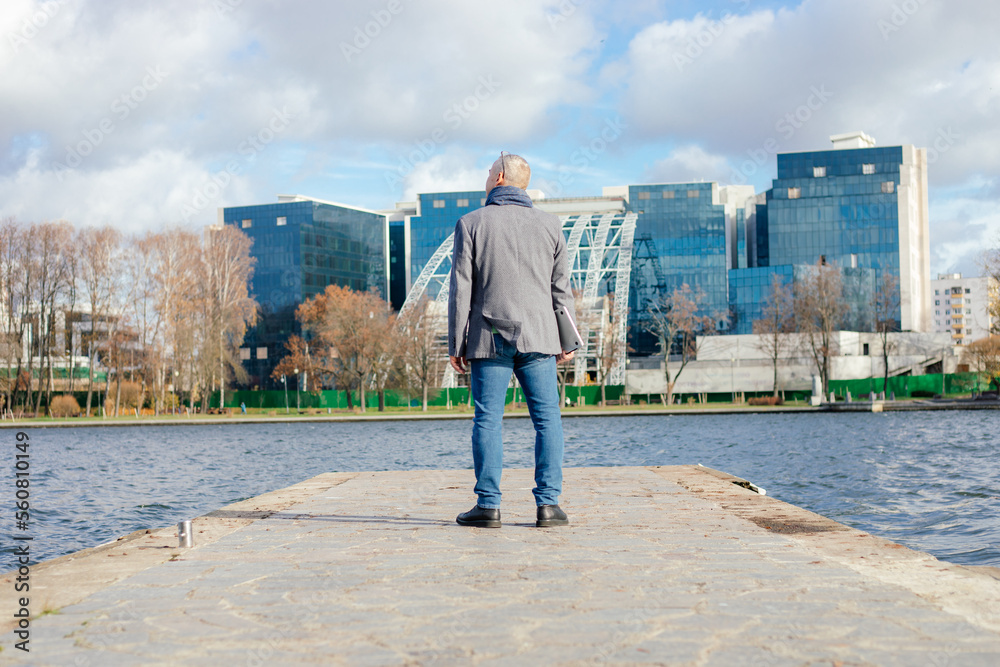 Rearview of gray haired businessman in warm jacket and scarf standing on river bridge,look office buildings. Urban area