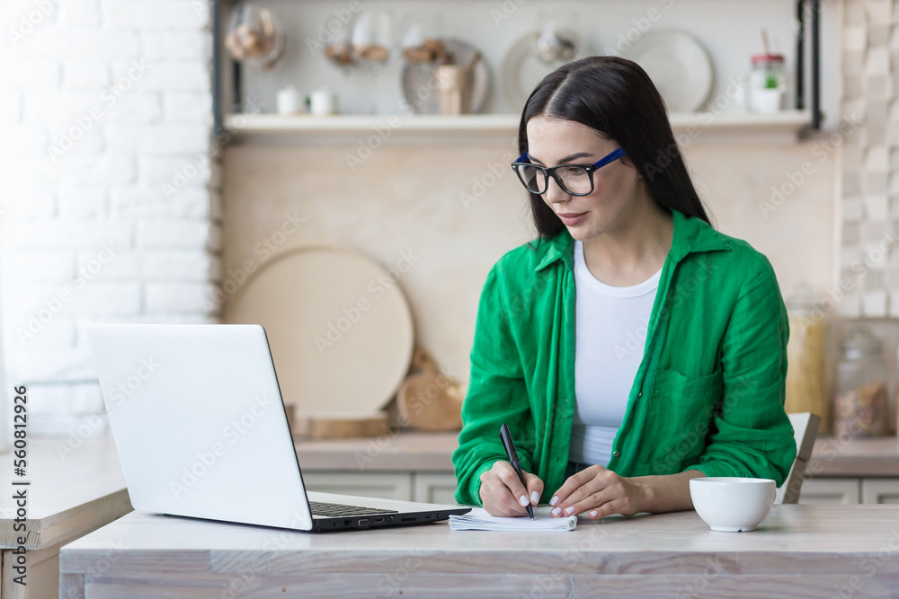 A young serious woman is having a remote job interview online. Sitting at home in the kitchen with a laptop. Speaks through a video call, introduces himself, passes the exam, writes in a notebook. A young serious woman is having a remote job interview online. Sitting at home in the kitchen with a laptop. Speaks through a video call, introduces himself, passes the exam, writes in a notebook.