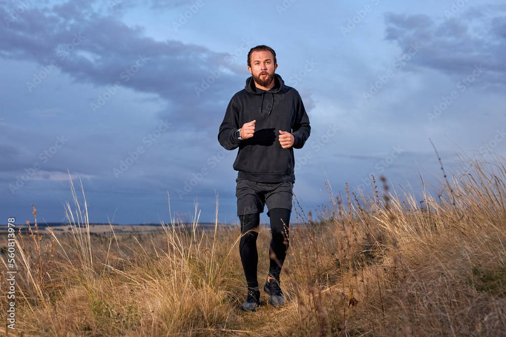 Jogger Young Caucasian Male Running Through the field. Man Wearing Black sportive wear. athlete guy with beard is motivated to be muscular, strong, in perfect physical shape. sport, fitness