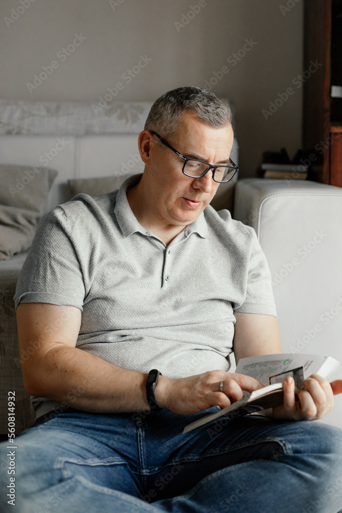 Vertical serious gray haired man in eyeglasses reading book, work in textbook, sitting on sofa indoors. Home education Vertical serious gray haired man in eyeglasses reading book, work in textbook, sitting on sofa indoors. Home education