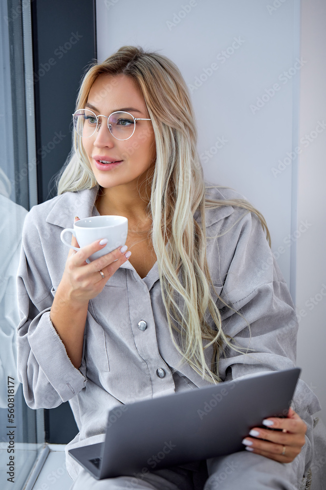Young beautiful female dressed casually working on laptop while sitting on window sill at home. Work from home at cozy atmosphere concept. good-looking female work and drink coffee. holding cup Young beautiful female dressed casually working on laptop while sitting on window sill at home. Work from home at cozy atmosphere concept. good-looking female work and drink coffee. holding cup