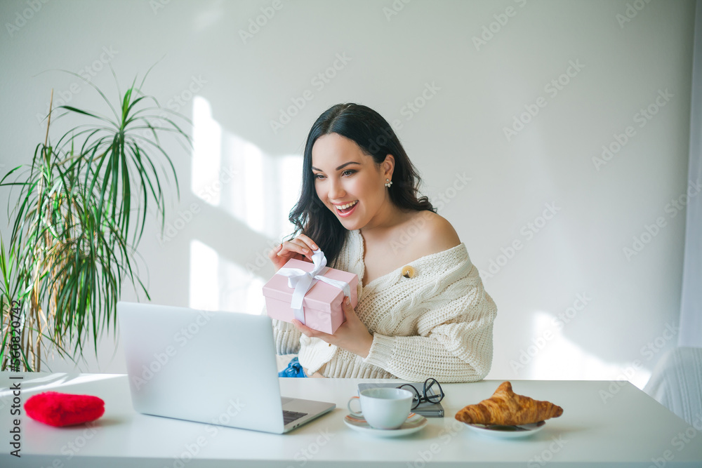 Young beautiful dark-haired woman of 34 years old in knitted sweater is talking on video link and holding pink gift box in her hands. Valentine's Day. Model received gift. Young beautiful dark-haired woman of 34 years old in knitted sweater is talking on video link and holding pink gift box in her hands. Valentine's Day. Model received gift.