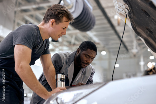 black and american or caucasian men can fix anything. handsome car mechanics in overalls uniform checking the engine under hood in modern clean car service station, workshop. team work