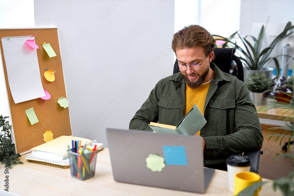 Smiling handsome office worker man sit reading plan, holding notebook in hands, bearded caucasian male in casual wear concentrated focused on work, generating business ideas. success, office Smiling handsome office worker man sit reading plan, holding notebook in hands, bearded caucasian male in casual wear concentrated focused on work, generating business ideas. success, office