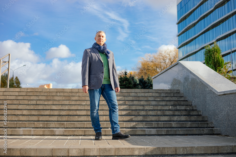 Serious mature grizzled businessman in jacket and scarf look camera, stand on staircase near office building. Promotion Serious mature grizzled businessman in jacket and scarf look camera, stand on staircase near office building. Promotion