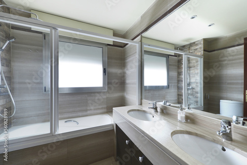 Corner of a toilet with a large mirror on the wall  a two-bowl sink in white porcelain and beige marble and a hydromassage unit with a glass partition