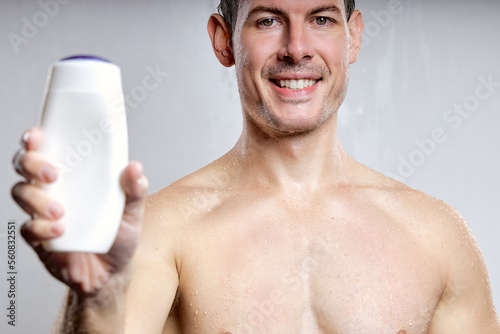 close-up portrait of handsome guy showing bottle of gel or shampoo, for advertisement, isolated on gray studio background. young male of caucasian appearance smiling, pleasant and nice, shirtless photo