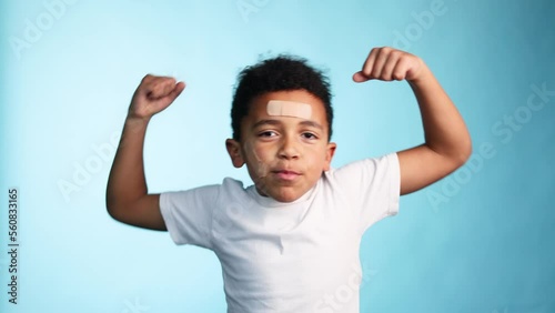 Portrait of clumsy brave smiling african american little boy with adhesive plaster looking at the camera and showing his muscles on isolated blue background Happy childhood  photo