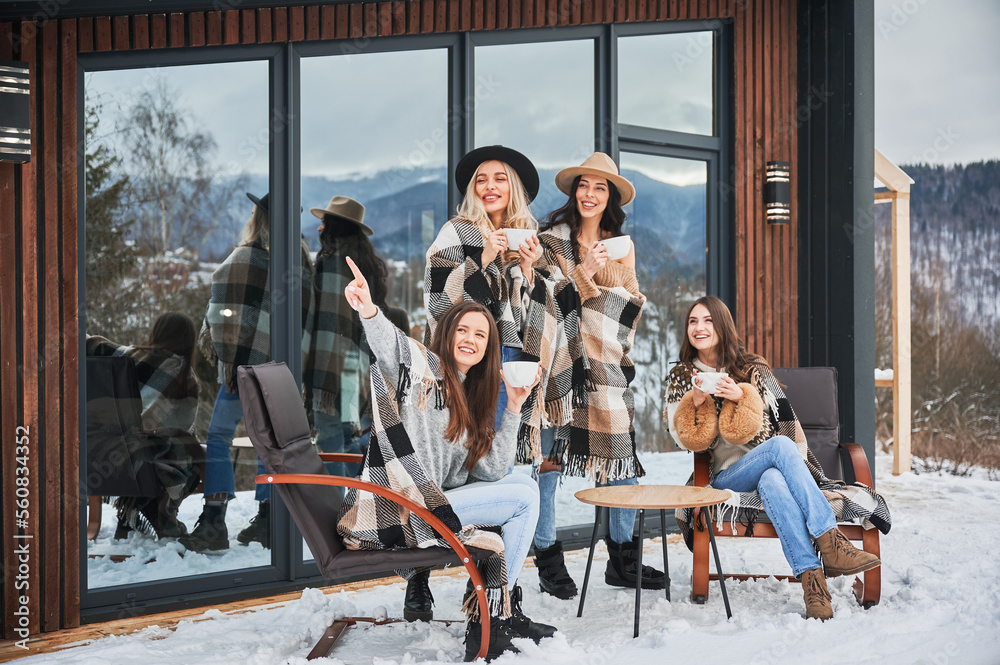 Young women enjoying winter weekends on terrace of contemporary barnhouse in the Scandinavian style. Four girls having fun, sitting on chairs and drinking hot tea.