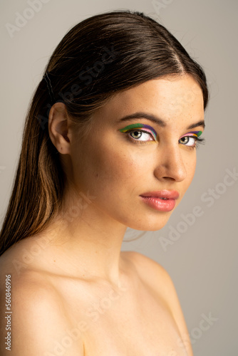 Teen model with naked shoulders and colorful eyeshadows looking at camera isolated on grey.
