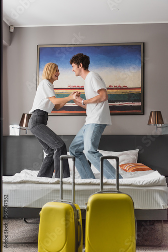 side view of excited couple holding hands and having fun on bed near baggage in modern hotel bedroom.