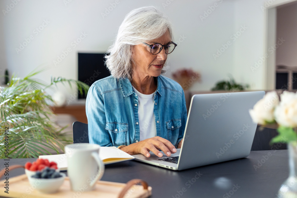 Shot of beautiful senior woman working with laptop while checking some documents in the kitchen at home. Shot of beautiful senior woman working with laptop while checking some documents in the kitchen at home.