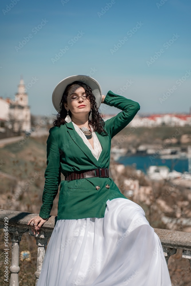 Woman walks around the city, lifestyle. A young beautiful woman in a green jacket, white skirt and hat is sitting on a white fence with balusters overlooking the sea bay and the city.