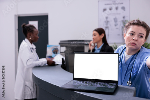 Laptop computer with white isolated screen standing on hospital counter desk during checkup visit appointment. Physician doctor discussing medical expertise with receptionist, medicine concept