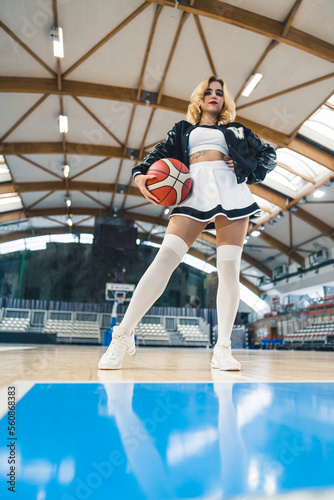 An attractive cheerleader is captured confidently holding a basketball, standing in an indoor basketball arena. She is dressed in a playful cheerleading outfit, with one hand resting on her waist.