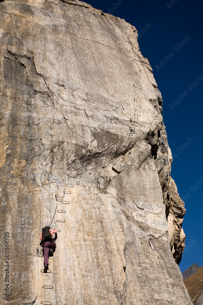 A young woman climbs up a ladder embedded in the cliff while engaging in the sport of Via Ferrata in Val D'isere, Savoie, France A young woman climbs up a ladder embedded in the cliff while engaging in the sport of Via Ferrata in Val D'isere, Savoie, France