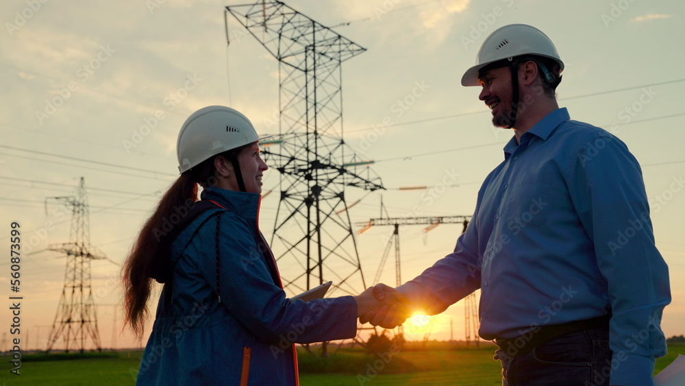 Two construction engineers work together on an electrical transmission line. Teamwork of power engineers in protective helmets, maintenance of power lines in outdoors. Colleagues shake hands in sun.