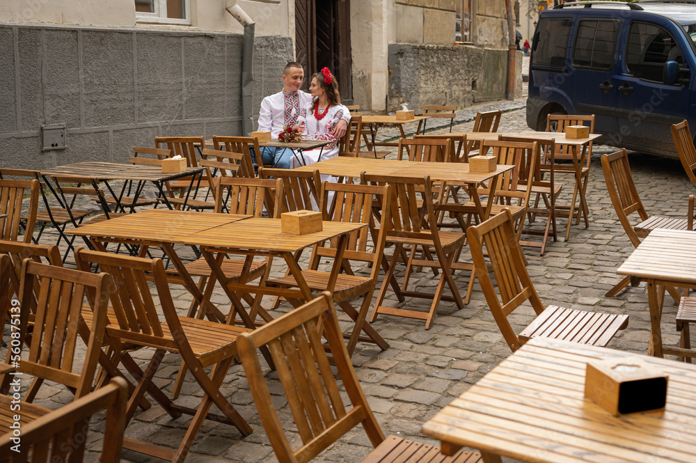 Fototapeta premium a young couple in Ukrainian national clothes, an embroidered woman sitting at a wooden table on the street, a terrace in a cafe