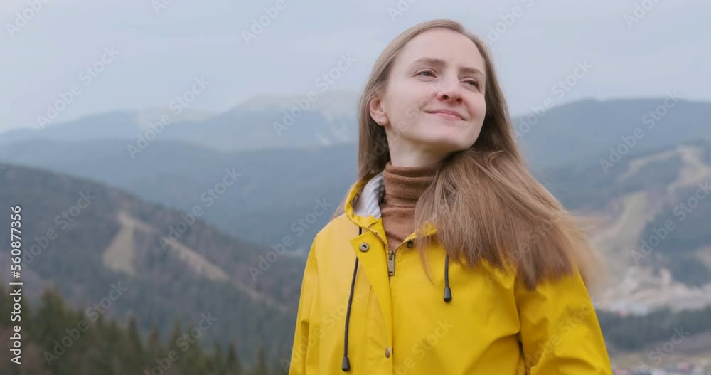 Happy woman adjusts hair and stretches arms top of cliff. Girl in yellow jacket on forest and mountains background Happy woman adjusts hair and stretches arms top of cliff. Girl in yellow jacket on forest and mountains background