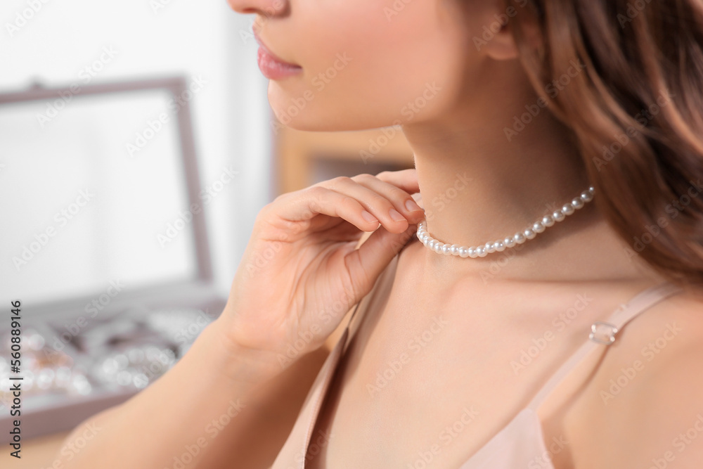 Young woman trying on elegant pearl necklace indoors, closeup