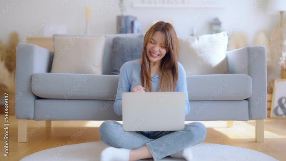 Young asian woman in good spirits working on laptop at home while sitting on the floor close to the couch. Excited female winner celebrating success while glancing at notebook Young asian woman in good spirits working on laptop at home while sitting on the floor close to the couch. Excited female winner celebrating success while glancing at notebook