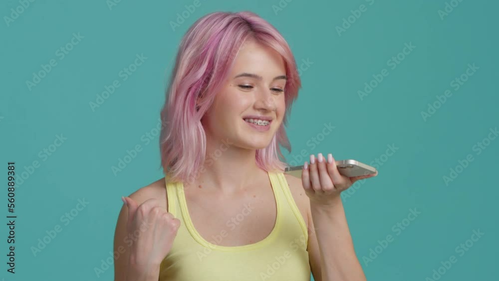 Smiling young woman 20s pink hair recording voice message on speakerphone. Student using virtual assistant app sets reminder on smart phone at studio background. Digital AI mobile assistance tech shot Smiling young woman 20s pink hair recording voice message on speakerphone. Student using virtual assistant app sets reminder on smart phone at studio background. Digital AI mobile assistance tech shot