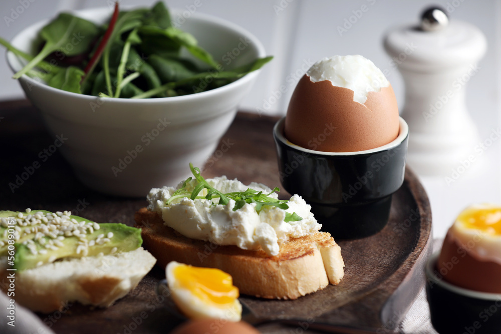 Fresh soft boiled egg in cup and sandwiches on wooden tray