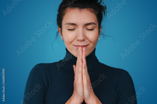 Positive cheerful woman keeping hands in praying gesture, posing against blue background. Woman believes in better, belief, faith. Close up portrait